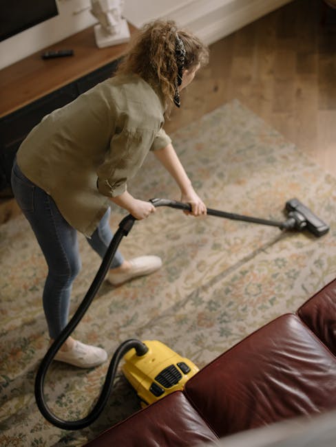 A woman with curly hair, wearing a beige jacket, dark jeans, and white shoes, is using a yellow vacuum cleaner with a black hose to perform surface cleaning on a patterned area rug in a living room. The room features wooden flooring partially covered by the rug, a wooden sideboard with a white decorative object, and a brown leather sofa visible in the foreground. The area is well-lit, highlighting the cleanliness and maintained condition of the surfaces. This image relates to domestic cleaning services offered by Carpet Cleaning Maida Vale, emphasizing deep cleaning and sanitisation suitable for booking in Maida Vale via their website.