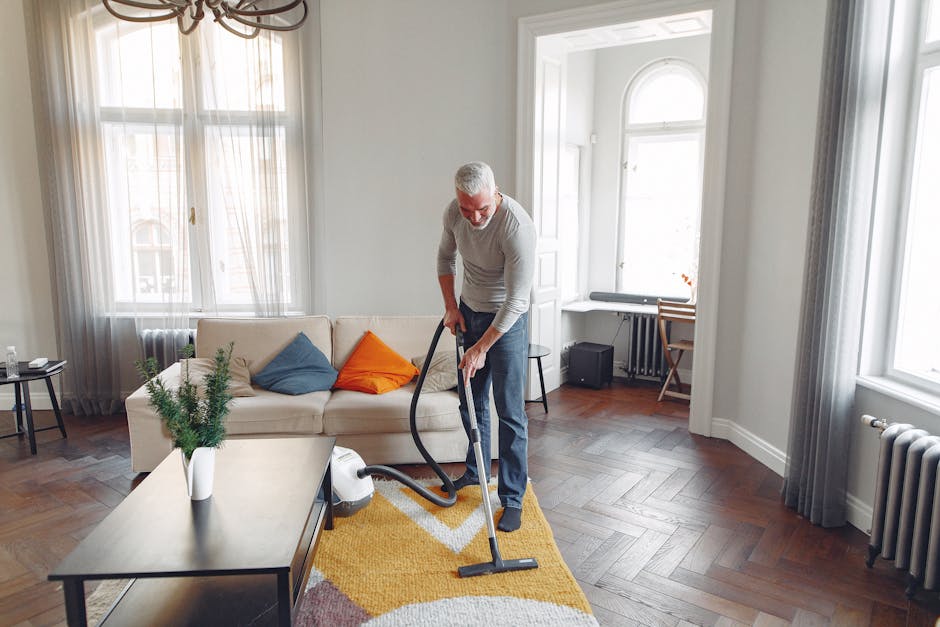 An elderly man with white hair and a beard is vacuuming a patterned yellow, white, and pink area rug in a bright, well-lit living room. The room features large windows with sheer white curtains, wooden flooring, a white sofa adorned with a blue and an orange cushion, and a black side table. There is a small potted plant on a coffee table and a desk with a chair near the windows. The man is dressed in a light gray long-sleeve shirt and dark pants, using a handheld vacuum cleaner to thoroughly clean the rug, reflecting a focus on surface cleaning and maintenance. Carpet Cleaning Maida Vale specializes in domestic cleaning services aimed at maintaining hygiene and freshness in residential spaces.