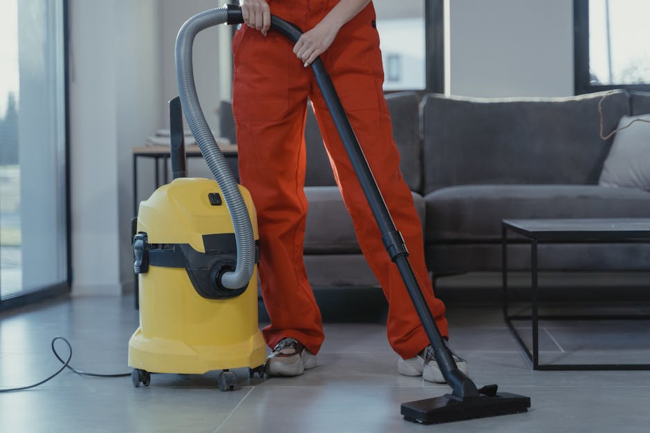 A person in orange work trousers and grey shoes is using a yellow vacuum cleaner with a black hose to clean the tile floor of a modern lounge area. The vacuum cleaner is positioned on the light grey t