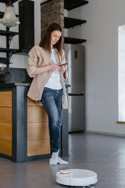 A person in orange work trousers and grey shoes is using a yellow vacuum cleaner with a black hose to clean the tile floor of a modern lounge area. The vacuum cleaner is positioned on the light grey t