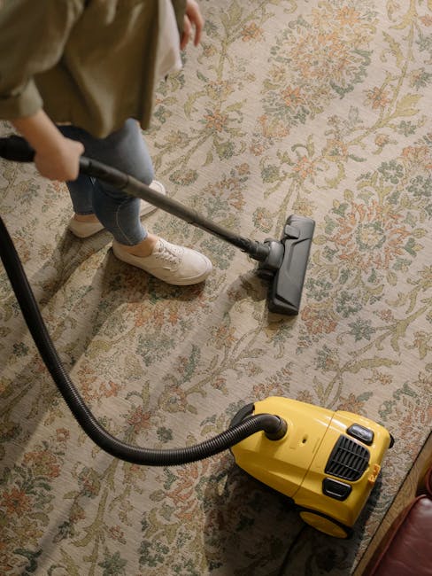 A person standing on a light grey tiled floor in a modern living room, holding a vacuum cleaner with a white and grey body and a long flexible hose, preparing for surface cleaning or deep cleaning as 