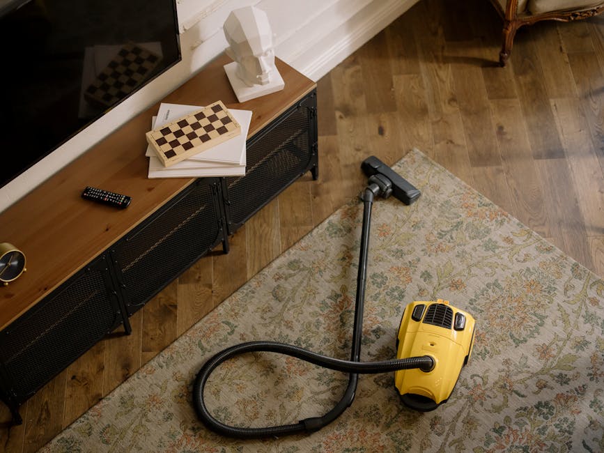A woman in casual clothing is vacuuming a large, patterned area rug with ornate floral designs in shades of beige, green, and orange. The rug is placed on a wooden floor inside a living room, next to