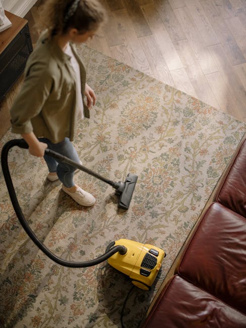 A person using a yellow vacuum cleaner to deep clean a patterned, beige and light green area rug in a living room. The individual is dressed in casual clothing, including a beige jacket and jeans, and is standing on a wooden floor adjacent to the rug. The room features a brown leather sofa and has ample natural lighting, highlighting the cleanliness of the surface. The scene demonstrates surface cleaning and maintenance as part of domestic cleaning services provided by Carpet Cleaning Maida Vale, focusing on hygiene and thorough dirt removal.