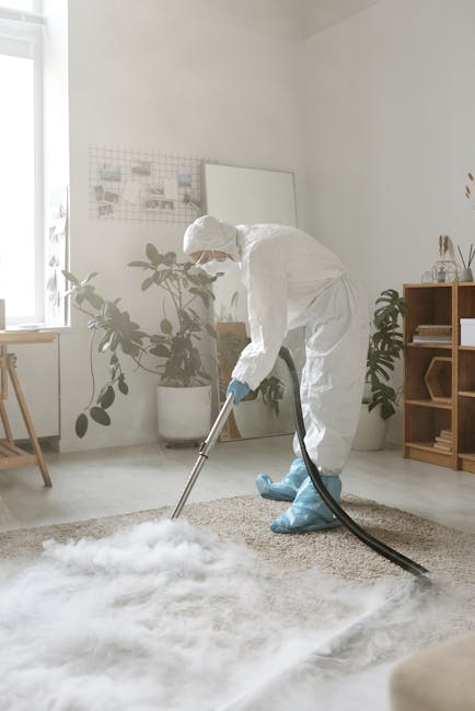 A professional cleaner dressed in full protective gear, including a white suit, face mask, gloves, and goggles, using a steam or fogging device to sanitise a carpeted area in a modern living room. The room features a light-colored carpet, a wooden shelving unit with decorative items, potted plants, and soft natural lighting. The cleaning process appears thorough, focusing on deep cleaning and surface sanitisation, aligned with services offered by Carpet Cleaning Maida Vale for domestic and commercial spaces.